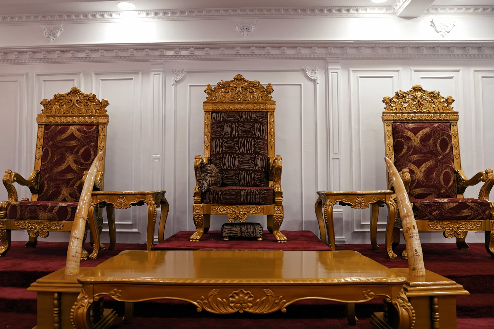 Three ceremonial palace thrones arranged beneath ornate gold detailing, with ivory tusks framing the central seat.