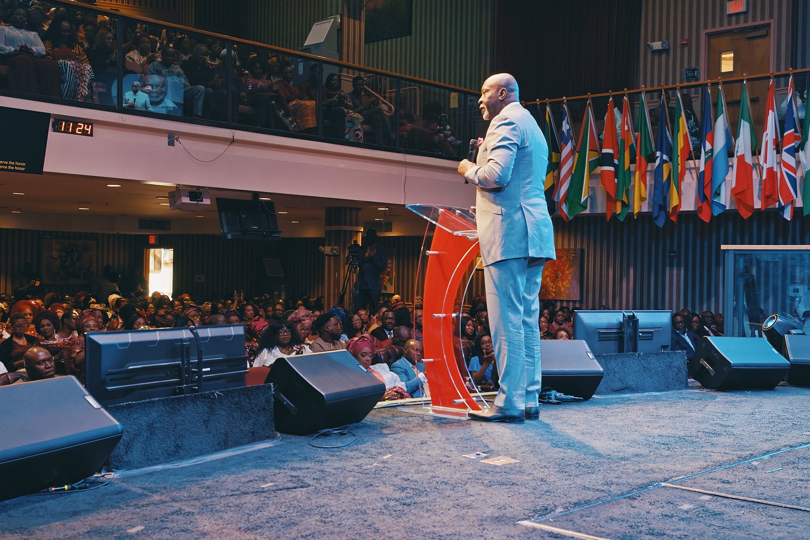 Ghandi Olaoye speaking on stage before a seated audience, with international flags behind him.