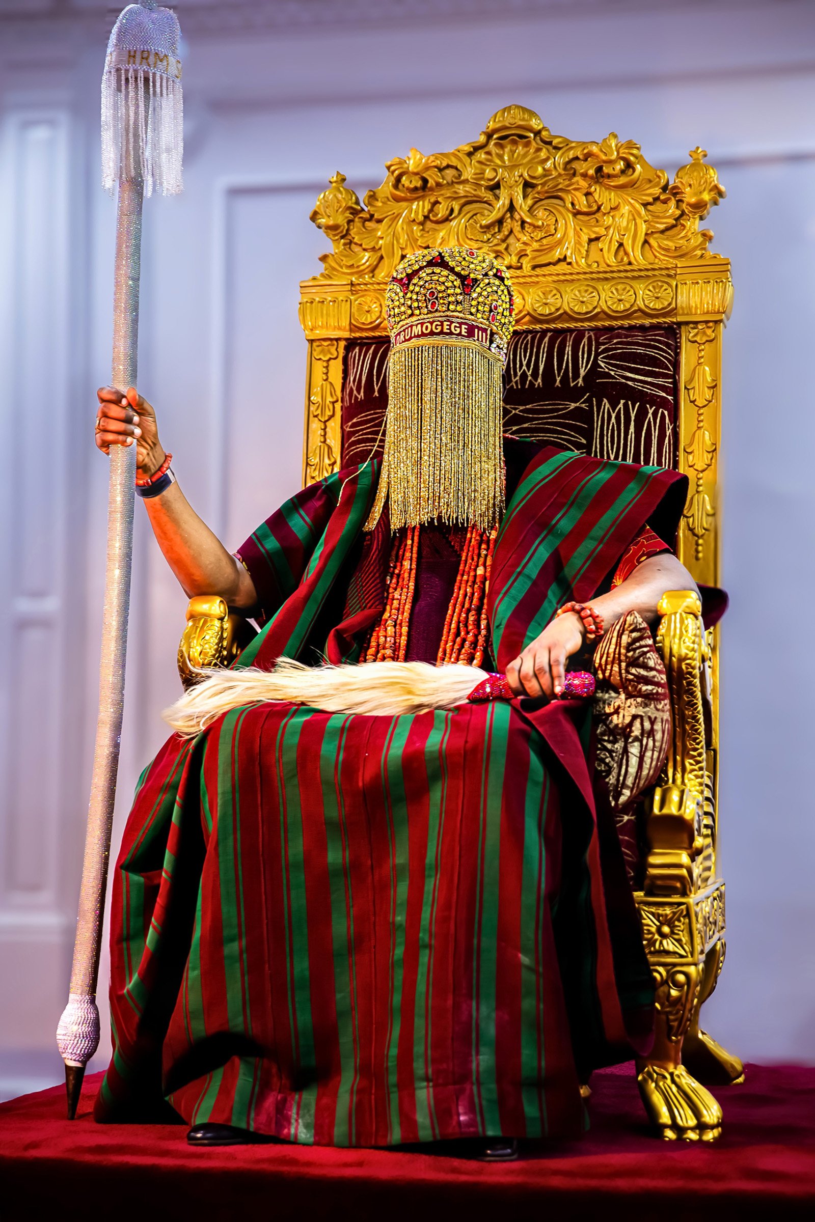 Oba Ghandi Olaoye seated on a gilded throne in red-and-green ceremonial robes, holding a silver staff.